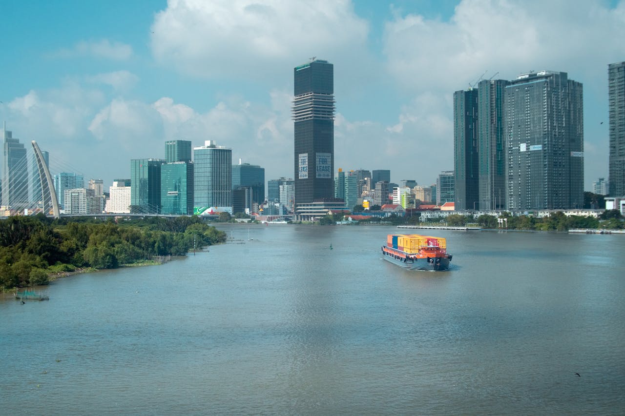 A cargo ship on a river against a city skyline with skyscrapers under a blue sky.